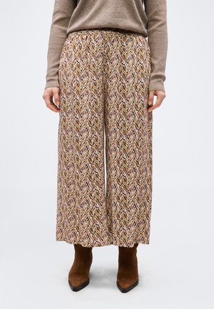 Žena nosi nohavice s širokými nohavicami s hnedým, ružovým a čiernym cikcak vzorom, v kombinácii s hnedými členkovými čižmami s špicatou špičkou a béžovým svetrom.