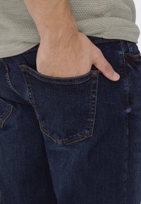 Mörkblå denimjeans med en lätt glans, som har en bakficka och kontrasterande orange sömmar. Hand delvis synlig i fickan.