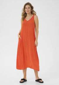 Orange ärmlös maxiklänning med V-ringning, svävande design, sidofickor och löst sittande passform. Stylad med svarta sandaler.