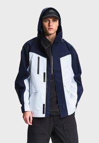 Jeune homme portant une veste à capuche bleu marine et blanche, un t-shirt noir avec des coutures blanches, un pantalon cargo noir et une casquette noire avec le texte "AIGLE".