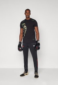 T-shirt noir avec un graphisme camouflage et une marque verte, associé à un pantalon de survêtement noir avec des touches rouges et des chaussures noires et beiges.