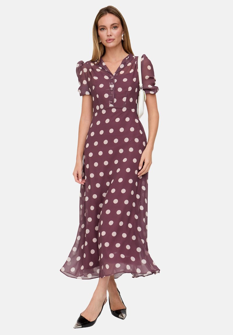 Das Kleid besteht aus einem weichen, fließenden Stoff in Burgunderrot mit großen weißen Punkten, hat eine durchgehende Knopfleiste und kurze, aufgebauschte Ärmel.