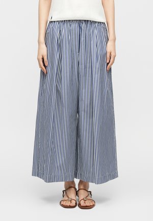 Nő kék-fehér függőleges csíkos bő szárú nadrágban, fehér felsőben és barna szandálban egyszerű háttér előtt.