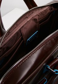 Intérieur d'un sac à main en cuir marron avec plusieurs poches, un compartiment zippé et des accents bleus contrastants. Texture lisse avec fermeture éclair métallique.