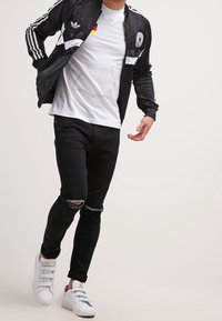 Giacca atletica nera con strisce bianche ed emblema della Germania, indossata sopra una maglietta bianca. Jeans neri strappati e sneakers bianche completano l'outfit.