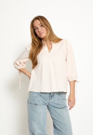 Žena s dlhými blond vlasmi, oblečená v svetloružovej blúzke s tromi štvrtinovými rukávmi a modrých džínsach, pózujúca na bielom pozadí.