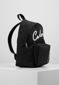 Czarny plecak o zaokrąglonym kształcie, z wyraźnym białym logo, zapinaną na zamek główną komorą oraz przednią kieszenią zapinaną na zamek.