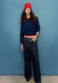 Maglione corto a righe navy con accenti rossi, abbinato a jeans a gamba larga in denim scuro. Il modello indossa un berretto lavorato a maglia rosso brillante.