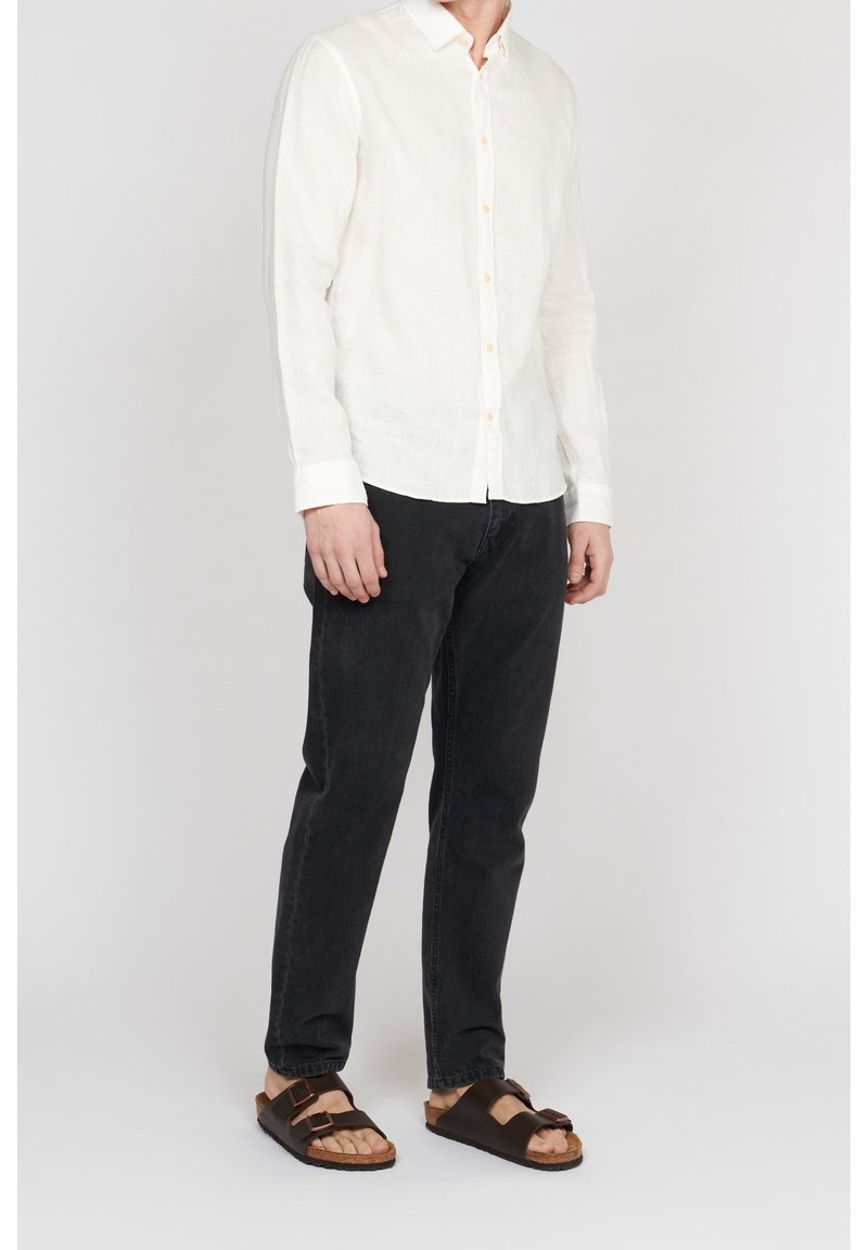 Homme portant une chemise blanche à manches longues boutonnée, un pantalon noir et des sandales marron à double bride, debout devant un fond uni.