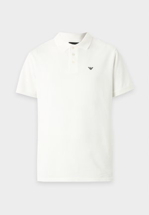 Uomo che indossa una polo bianca, pantaloncini beige, calzini neri, mocassini neri e una borsa a tracolla su sfondo bianco uniforme.