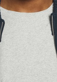 Närbild av en person som bär en ljusgrå t-shirt med rund halsringning och en delvis zipad mörkblå jacka med silverfärgade dragkedjetänder.