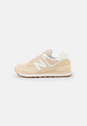 Beige og hvid New Balance-sneaker med ruskindsupperdel, snørelukning og riflet gummisål, vist i siden set mod en hvid baggrund.