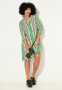 Multikleuren gestreepte jurk met groene, gele en rode accenten, 3/4 mouwen en een franjeszoom. Gecombineerd met zwarte platformklompen en een gestructureerde clutch.
