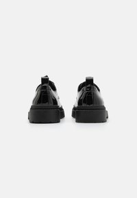 Zapatos de charol negros con una suela gruesa y texturizada, que presentan un tacón bajo y una lengüeta en la parte posterior para facilitar su calzado.