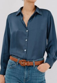Camicia di satin blu navy con maniche lunghe e colletto, con chiusura a bottoni. Indossata con jeans azzurri chiari e una cintura marrone.