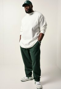 Camicia bianca a maniche lunghe, pantaloni verdi, sneakers bianche con accenti verdi. La persona indossa un cappello verde scuro e un orologio al polso.