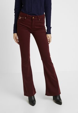 Vrouw draagt nauwsluitende donkerbordeauxrode uitlopende broek, donkerblauw shirt met lange mouwen en zwarte enkellaarzen, staand tegen een witte achtergrond.