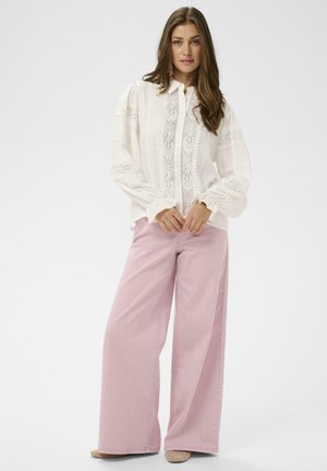 Kvinde iført en hvid bluse med lange ærmer og blonde detaljer samt højtaljede, vidde rosa bukser, stående foran en ensfarvet baggrund.