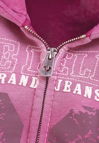 Veste à capuche zippée rose avec une fermeture éclair argentée. Présente un grand texte blanc "BELLA" et "GRAND JEANS". Tissu doux avec une surface légèrement texturée.