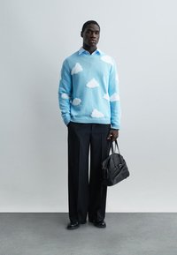 Άνδρας που φοράει μπλε πουλόβερ με λευκά σχέδια σύννεφων, μαύρο παντελόνι φαρδύ, μαύρα παπούτσια, κρατώντας μια μαύρη τσάντα, σε απλό γκρι φόντο.