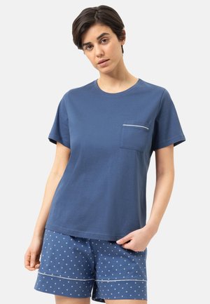 Femme portant un t-shirt bleu à manches courtes avec une poche poitrine et un short bleu à motifs avec une bordure blanche sur un fond clair uni.