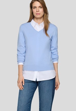 Frau mit langen hellbraunen Haaren, die einen hellblauen V-Ausschnitt-Pullover über einem weißen Hemd und blauen Jeans trägt, steht vor einem grauen Hintergrund.