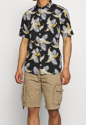 Chemise noire à manches courtes, à boutons, avec un imprimé floral et de feuilles de palmier jaunes et blanches ; associée à un short cargo beige avec des poches latérales.