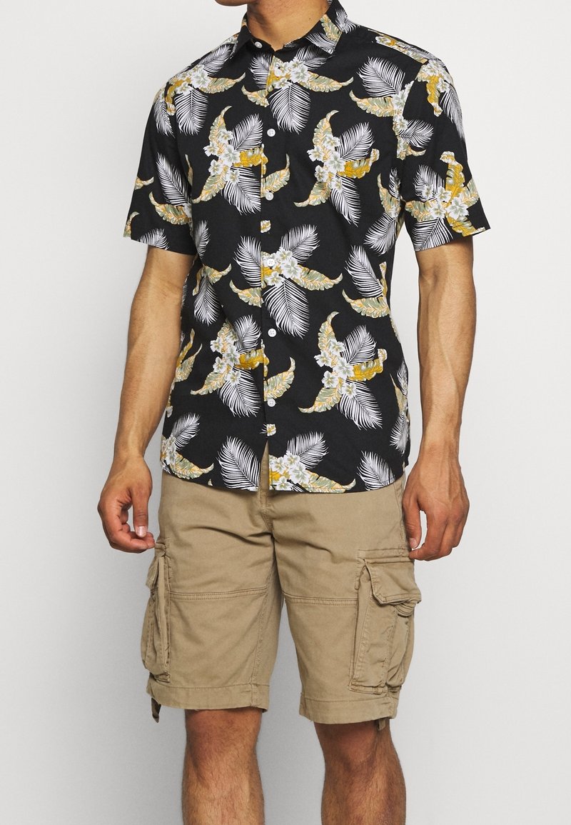 Korte mouwen, zwart overhemd met knopen en een gele en witte bloemen- en palmbladprint; gecombineerd met beige cargoshorts met zijzakken.