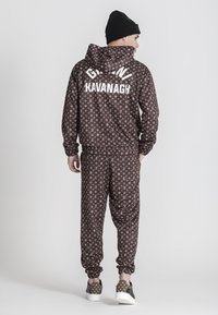 Chándal marrón con estampado y capucha, que presenta el texto blanco "GAVIN KAVANAGH". Combinado con zapatillas deportivas estampadas. Lleva un gorro de punto en la cabeza.