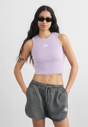 Νέα γυναίκα με μακριά σκούρα μαλλιά, φορώντας μαύρα γυαλιά ηλίου, λιλά κοντό τοπ και γκρι σορτς με κορδόνι, με τα χέρια στις τσέπες.