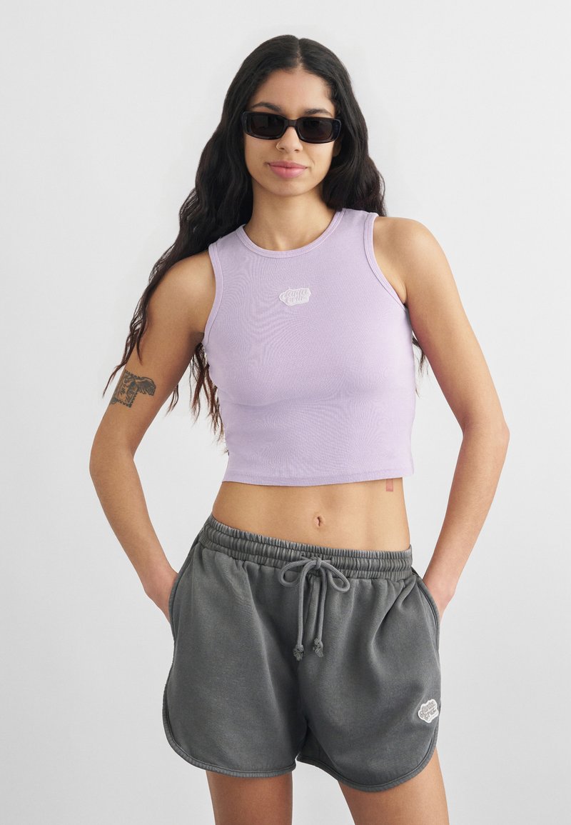 Νέα γυναίκα με μακριά σκούρα μαλλιά, φορώντας μαύρα γυαλιά ηλίου, λιλά κοντό τοπ και γκρι σορτς με κορδόνι, με τα χέρια στις τσέπες.