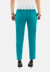 Pantalons fuselés teal avec poches latérales, présentant une texture lisse et des escarpins noirs contrastants. Au-dessus, un blouse en coton blanc.