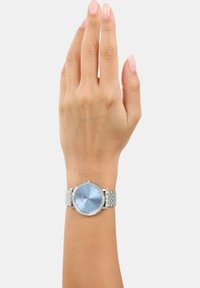 Silberne Armbanduhr mit einem runden blauen Zifferblatt, metallischem Armband und Strass-Akzenten um die Lünette. Zeiger sind so positioniert, dass die Uhr deutlich zu sehen ist.