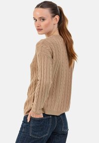 Beiger Strickpullover mit Kabelmuster, strukturiertem Design, Bündchen und rundem Halsausschnitt, kombiniert mit dunkelblauen Jeans.