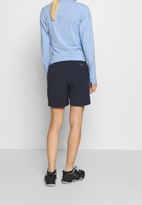 Žena v světle modrém dlouhém rukávu, černých šortkách a černých turistických botách stojící na jednobarevném bílém pozadí.