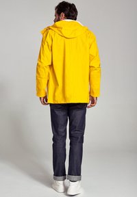 Gul vandtæt jakke med hætte, lange ærmer og sidelommer, parret med mørke jeans og hvide sneakers. Glat tekstur.
