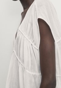 Haut en lin blanc à manches courtes, avec des fronces verticales et un décolleté en V. La texture lisse et le design léger favorisent la respirabilité.