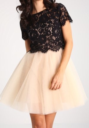 Frau trägt ein schwarzes Spitzenoberteil mit kurzen Ärmeln, kombiniert mit einem knielangen beigen Tüllrock.