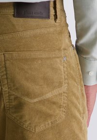 Braune Corduroy-Latzhosen verfügen über eine vertikal gerippte Textur, seitliche Taschen und ein Leder-Brandlabel. Der Stoff wirkt weich und strapazierfähig.