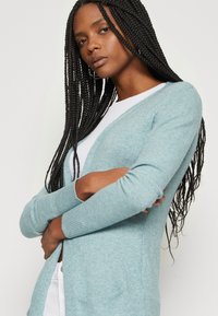 Lyseblå cardigan med en myk tekstur, åpen frontdesign og ribbede mansjetter. Modellen har lange fletter og armene krysset foran kroppen.