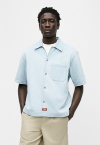 Νεαρός άνδρας που φοράει ανοιχτό γαλάζιο πουκάμισο με κοντά μανίκια πάνω από λευκό μπλουζάκι και μπεζ παντελόνι, στέκεται με τα χέρια στις τσέπες.