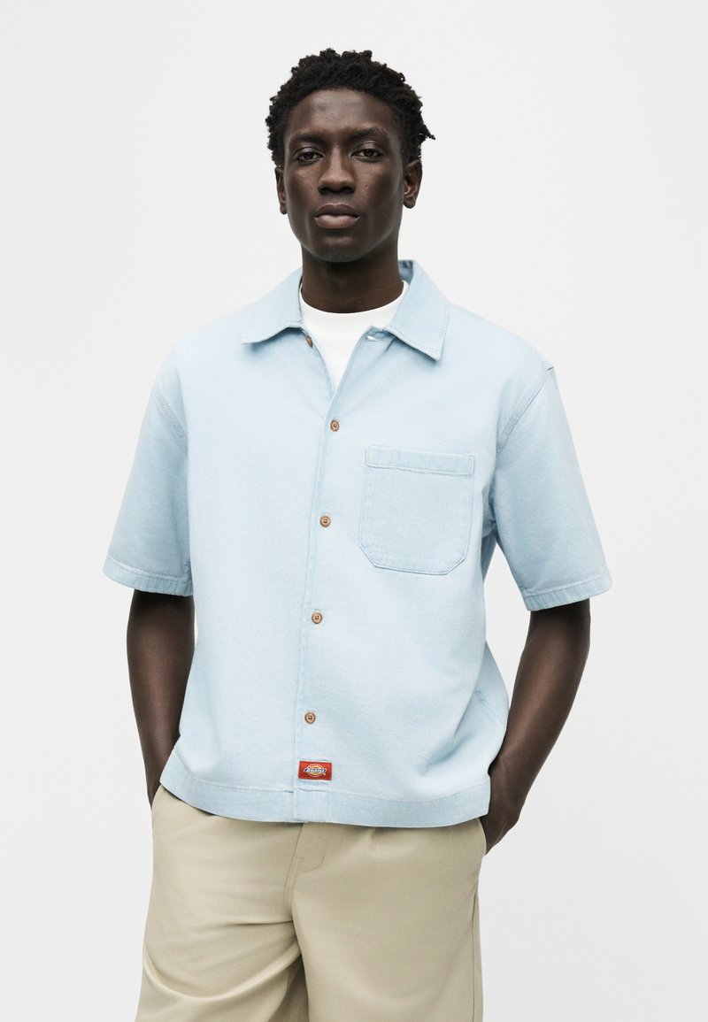 Jeune homme portant une chemise à manches courtes bleue claire par-dessus un t-shirt blanc et un pantalon beige, debout avec les mains dans les poches.