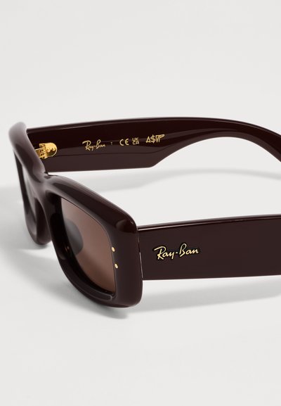 Тъмнокафяви слънчеви очила Ray-Ban с дебели правоъгълни рамки и златисто лого на дръжките и вътрешната страна на рамките.