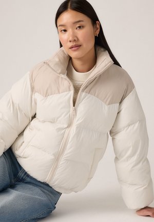 Puffy witte jas met een lichtbeige schouderstuk. Voorzien van een hoge kraag, een rits aan de voorkant en zijzakken. G gedragen over een crèmekleurige trui.