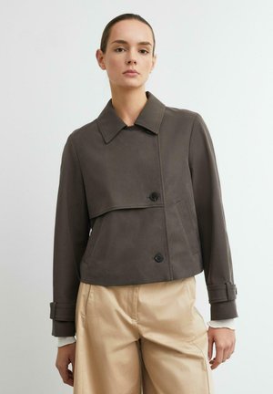 Frau trägt eine dunkelbraune kurze Jacke mit Knöpfen über beigen Hosen und steht vor einem schlichten hellen Hintergrund.