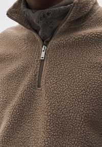 Pullover a mezza zip in pile marrone con una superficie testurizzata e zip argentata. Sotto, è visibile una sciarpa scura con motivo.