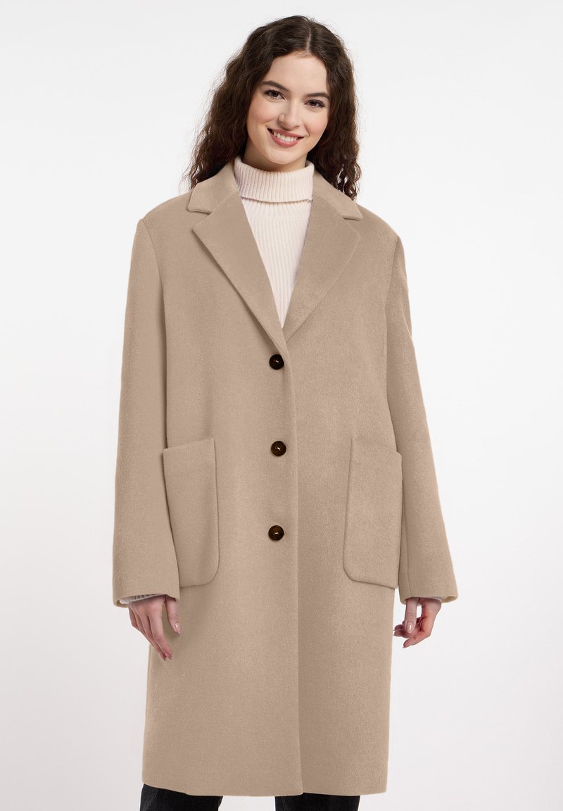 Beige wollen jas met een gekartelde kraag, twee grote voorkzakken en donkere knopen. Zachte textuur en middellange snit.