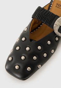 Black leather shoe featuring a square toe, elasticized strap, and silver-toned studs in a floral design across the upper surface.