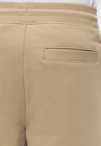 Gros plan sur la ceinture et la poche arrière d'un pantalon de survêtement beige avec un élastique côtelé et une poche fendue horizontale.