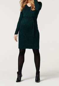 Zielona sukienka ciążowa w kwiatowy wzór, z dekoltem w serek, długimi rękawami, marszczoną talią i długością do kolan, zestawiona z czarnymi botkami za kostkę.